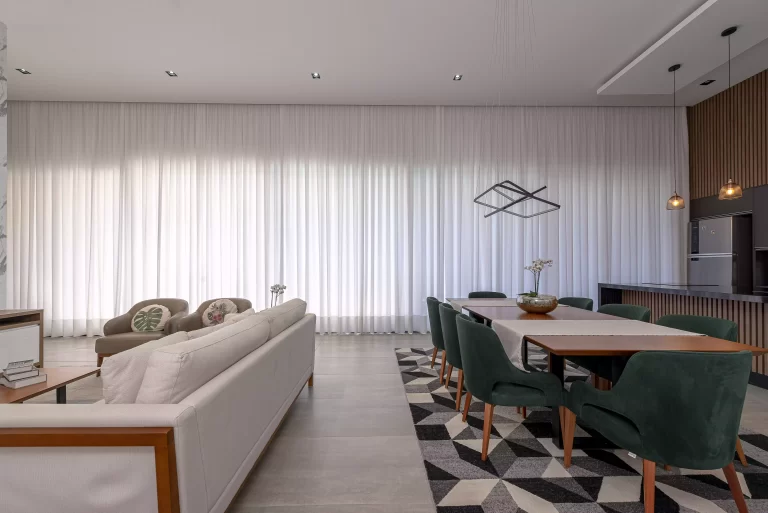 Sala de estar e de jantar com sofá à esqueda, uma mesa de jantar com cadeiras e tapete à direita, e ao fundo cortinas brancas wave